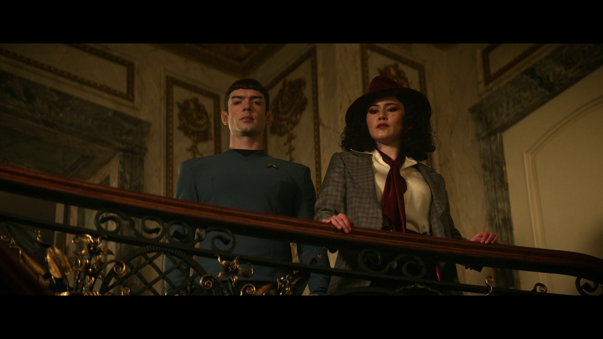 Spock and La'an people are standing on a decorative balcony, with Spock wearing his blue uniform and La'an in a checkered outfit and red hat which is very reminiscent of Carmen Sandiego.