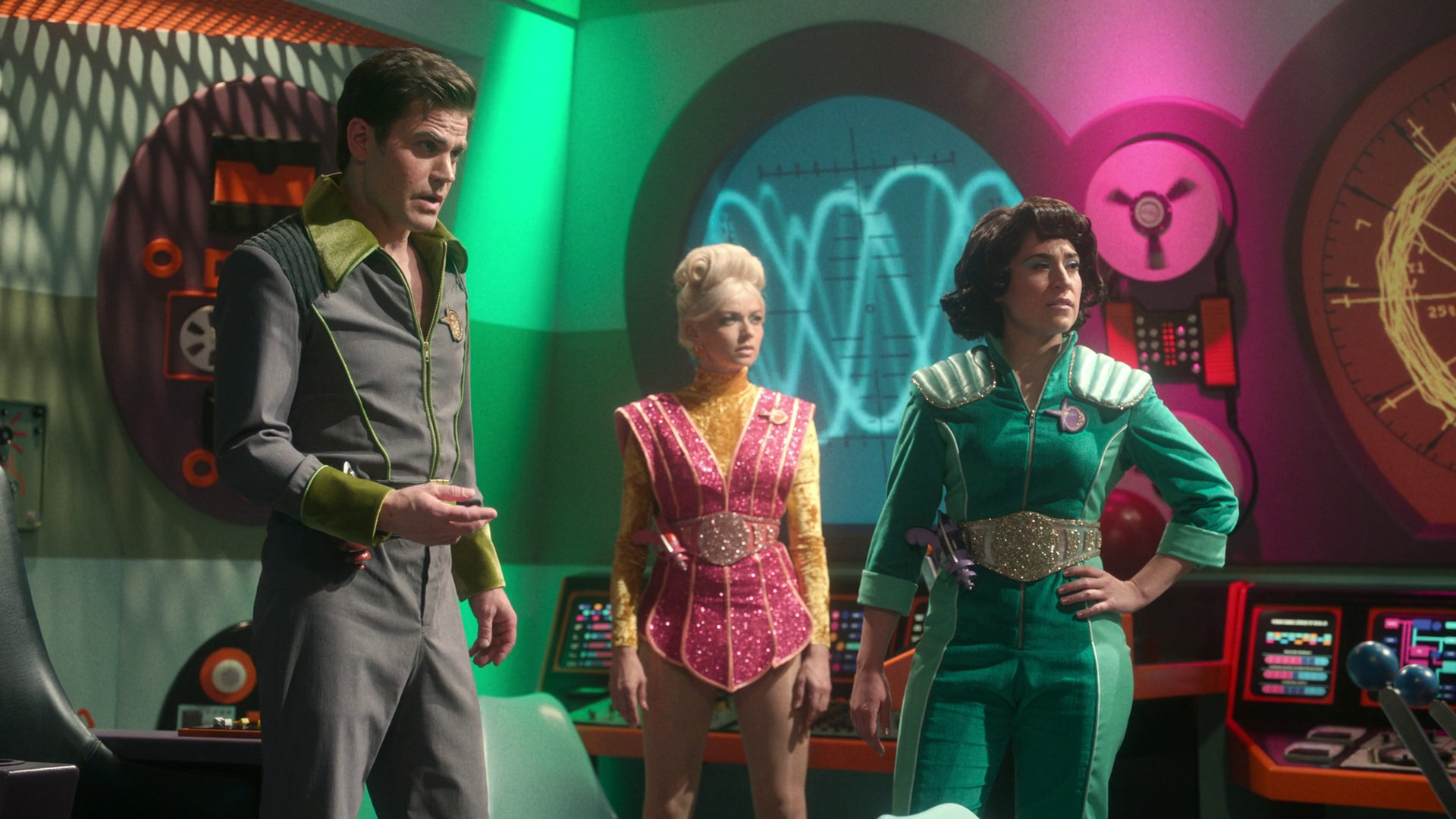 A retro-futuristic scene depicts three people wearing colorful, space-themed outfits standing in a room with vibrant, glowing control panels and walls.