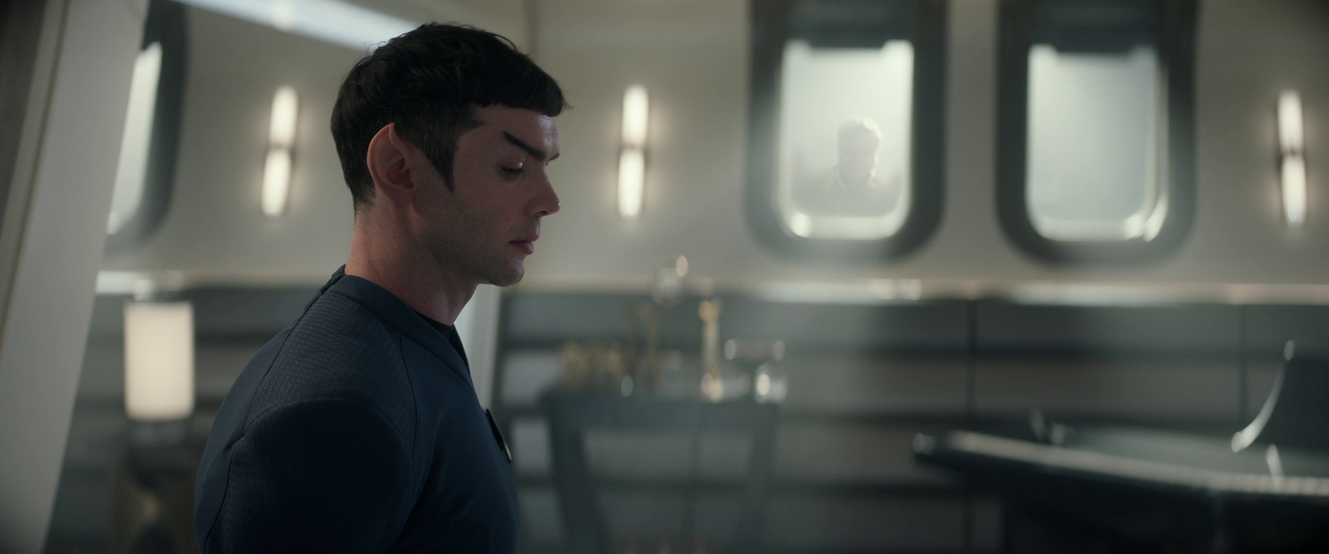 Spock stands in the foreground while a mysterious figure who is later revealed to be the cosmic entity Trelane watches from outside the window of his quarters.