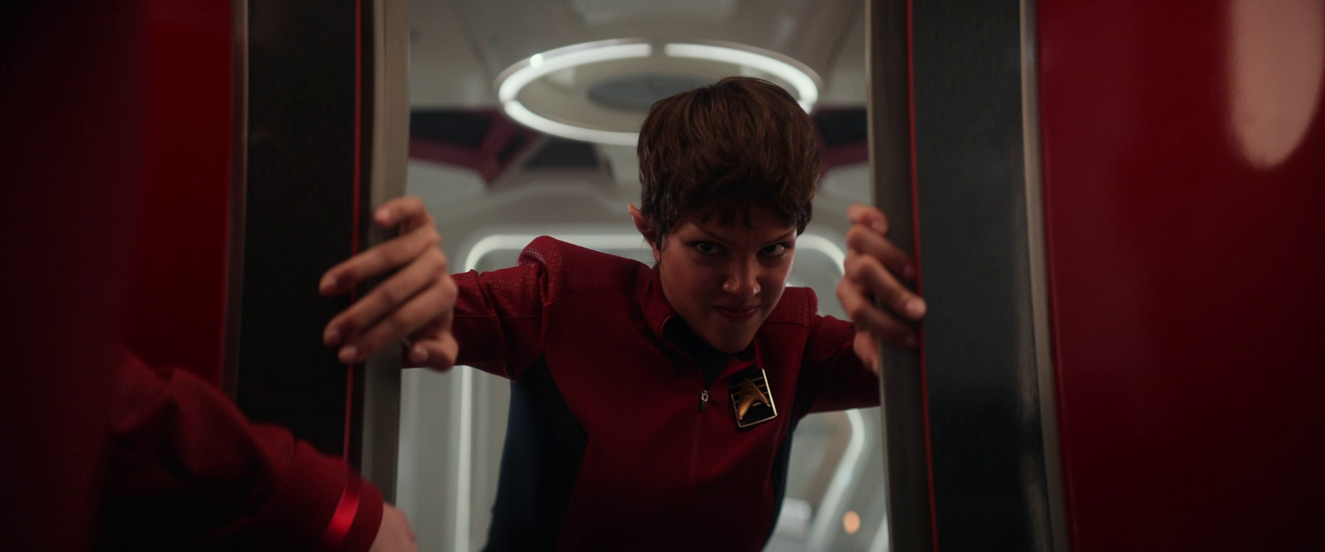 A person wearing a red uniform is gripping the edges of a doorway or hatch, looking intently forward in a futuristic or sci-fi setting.