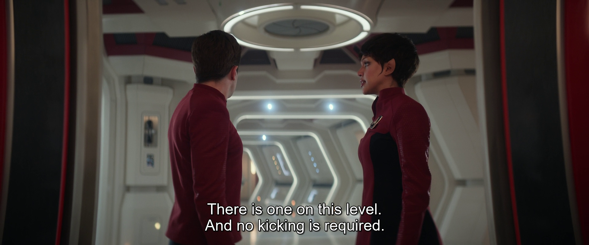 Two people in futuristic red uniforms are standing in a brightly lit corridor, having a conversation where one says, There is one on this level. And no kicking is required.