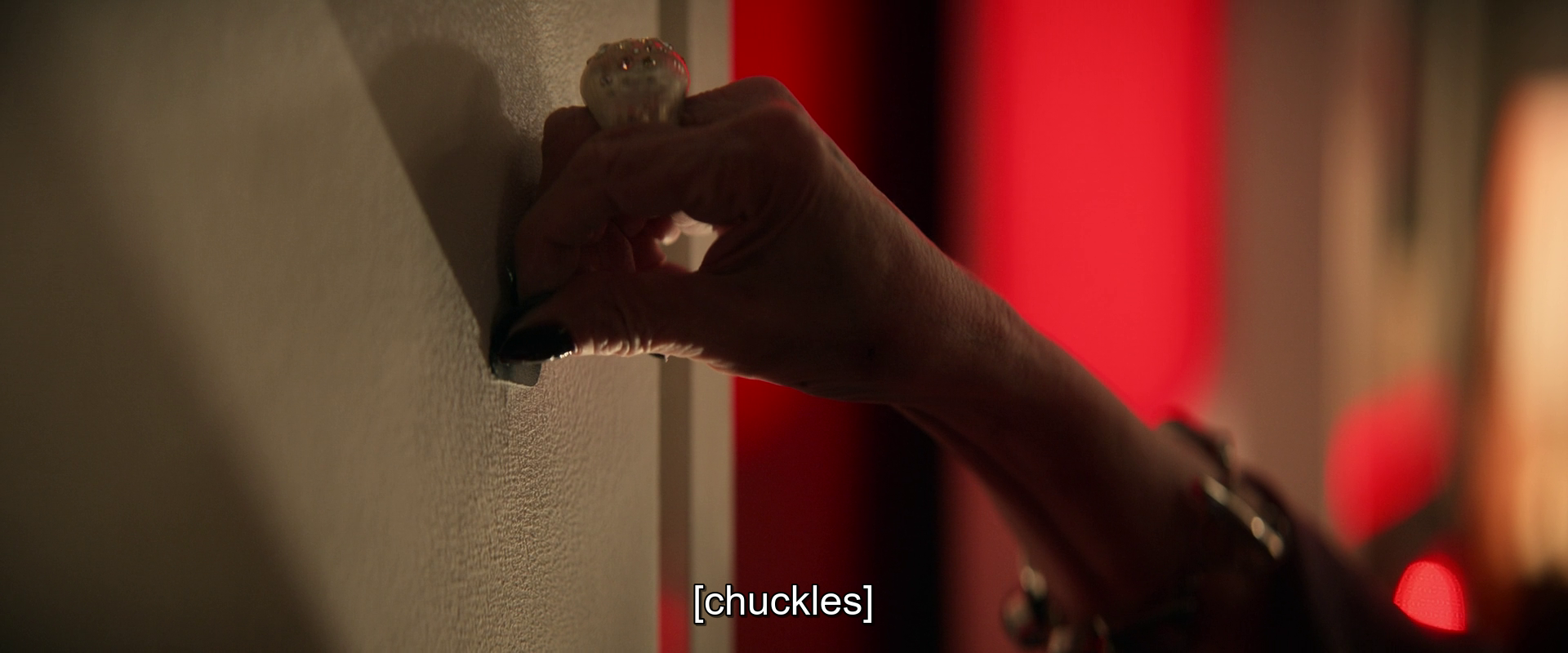 A hand adorned with multiple rings is turning a small knob or button on a wall, with a red light casting a glow in the background.