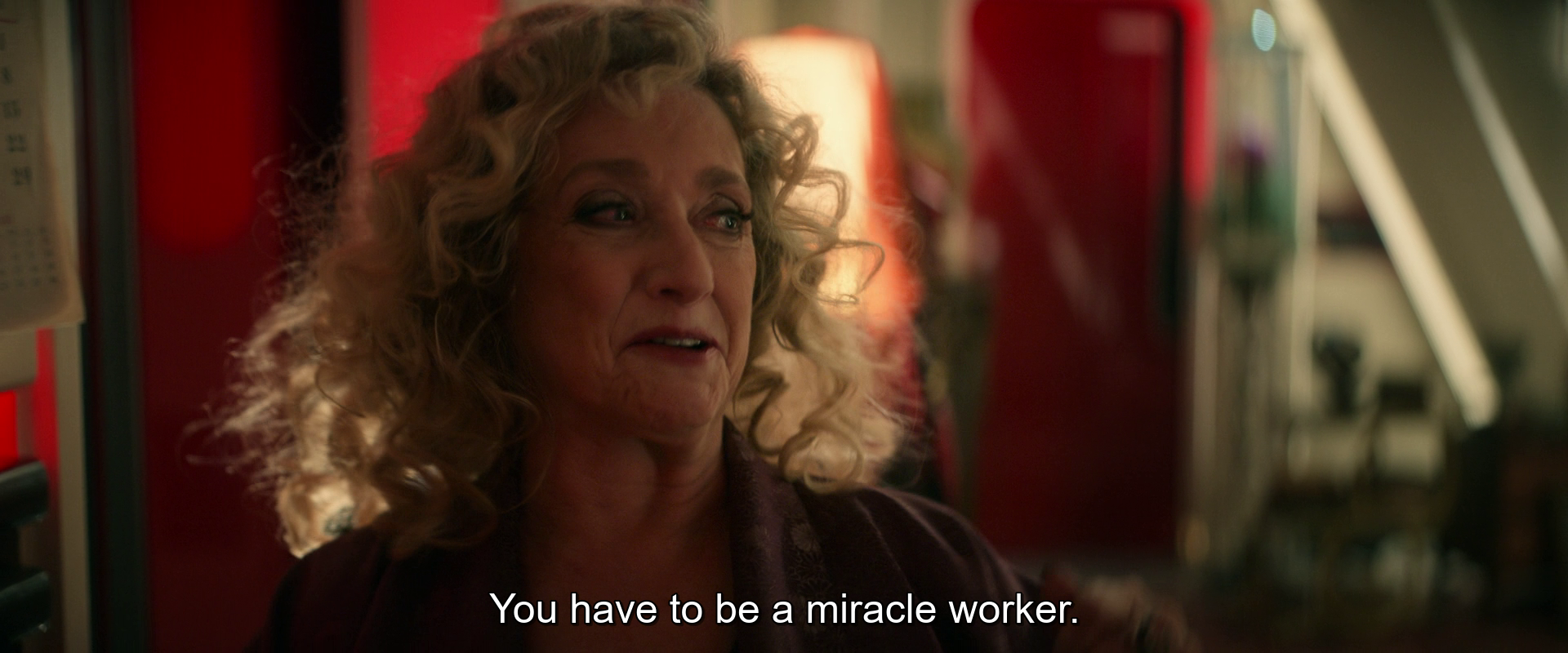 A woman with curly hair is speaking in a warmly lit room, saying, You have to be a miracle worker.