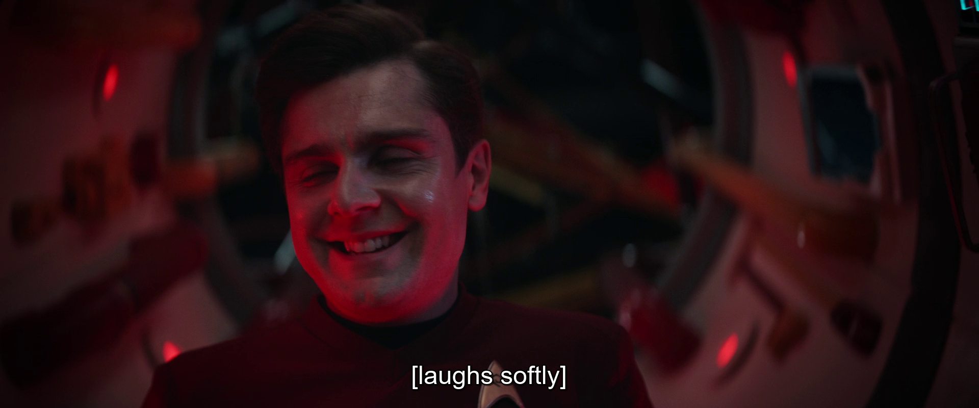 A man is softly laughing in a dimly lit, futuristic or sci-fi setting with a red hue.